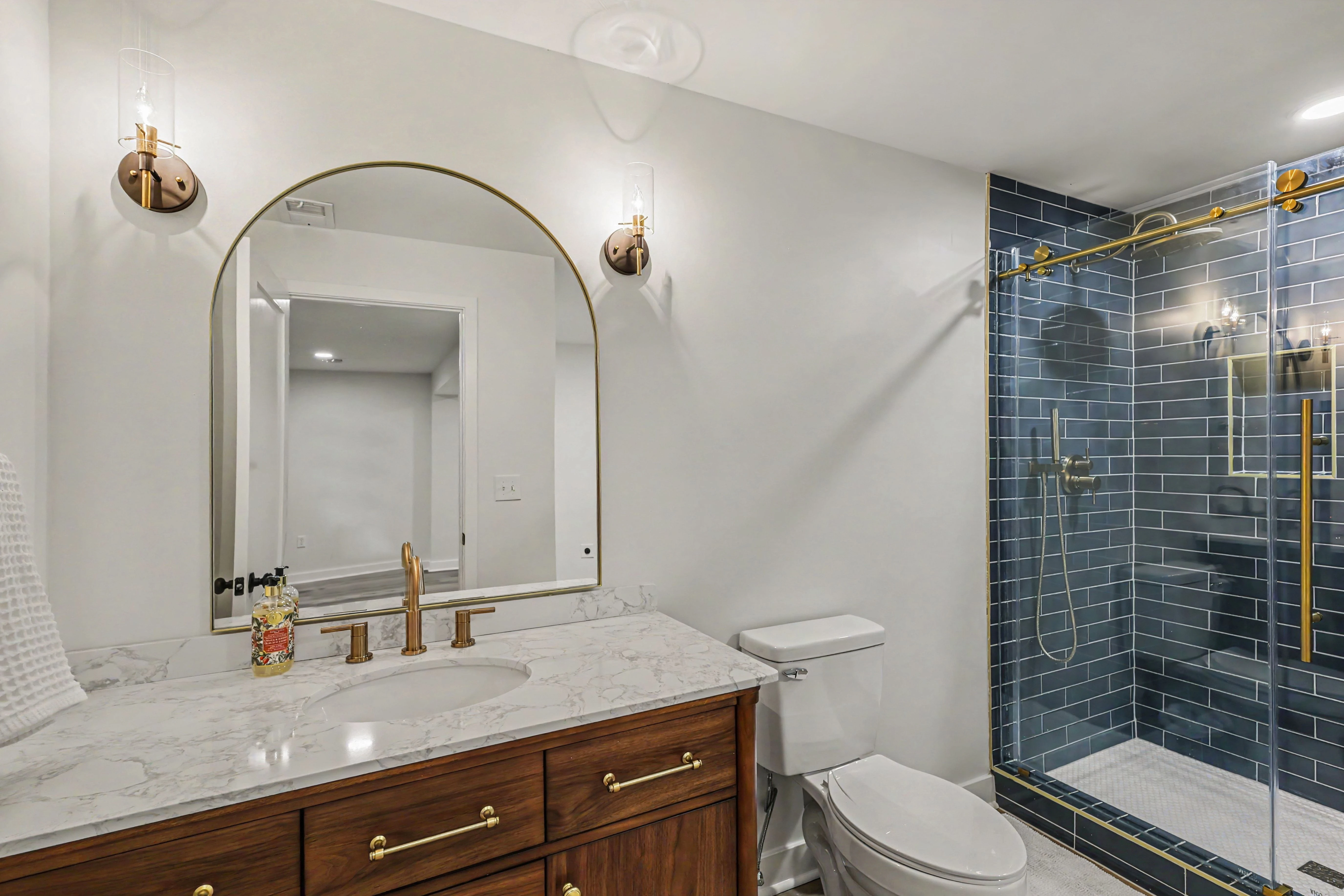 Basement bathroom with walnut vanity, arched brass mirror, and blue tiled shower in a Prairie Village basement remodel